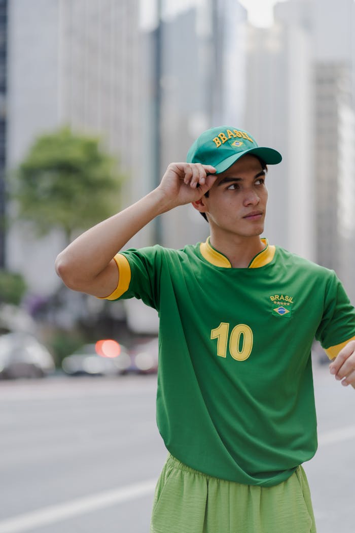 Young man wearing a green Brazilian soccer jersey and cap, standing on an urban street.