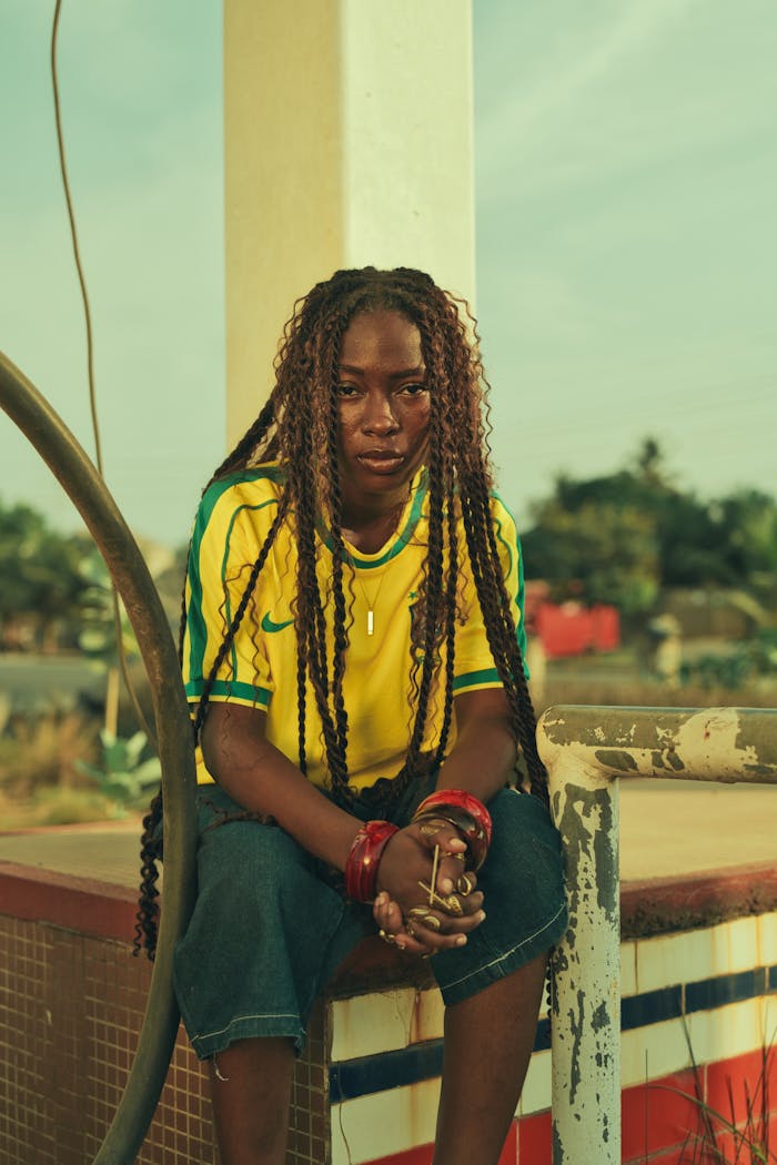Woman wearing Brazil soccer jersey with braids, sitting outdoors.